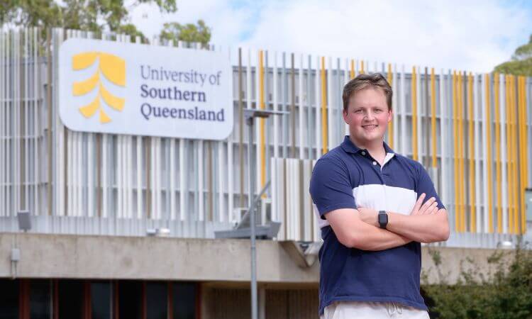 Person standing with arms crossed in front of the University of Southern Queensland building, wearing a navy and white polo shirt.