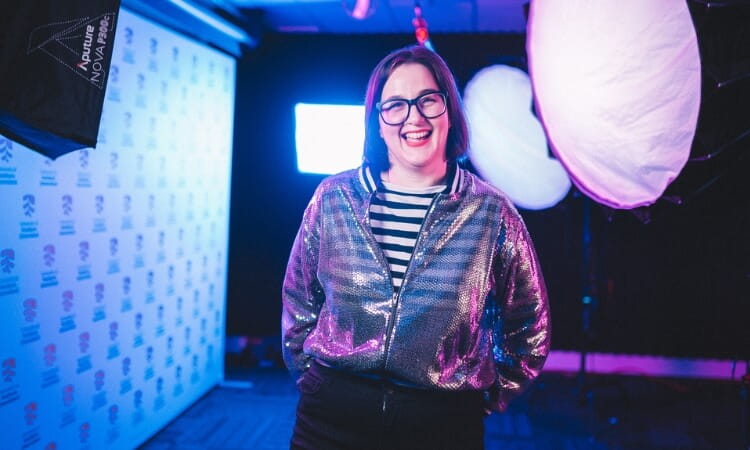 A person wearing glasses, a striped shirt, and a shiny silver jacket stands smiling in a brightly lit studio with photography equipment in the background.