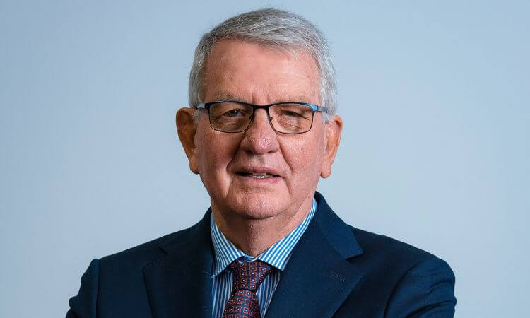An older man with short gray hair, glasses, and a suit with a striped shirt and patterned tie, stands against a plain light background.
