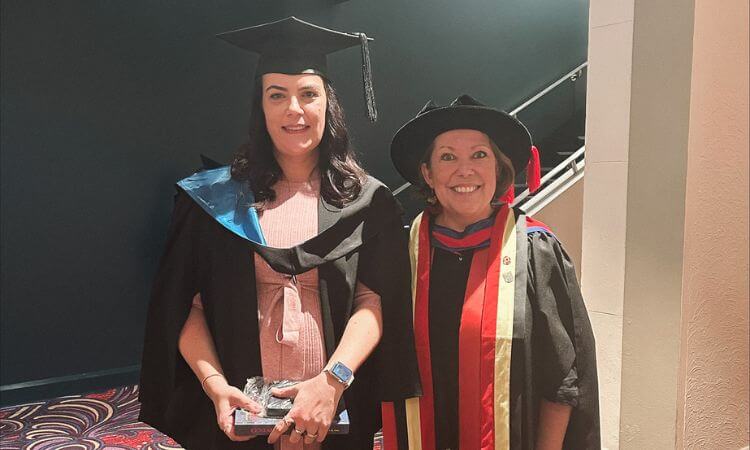 Two people in academic regalia at a graduation ceremony. One holds a plaque and both are smiling.