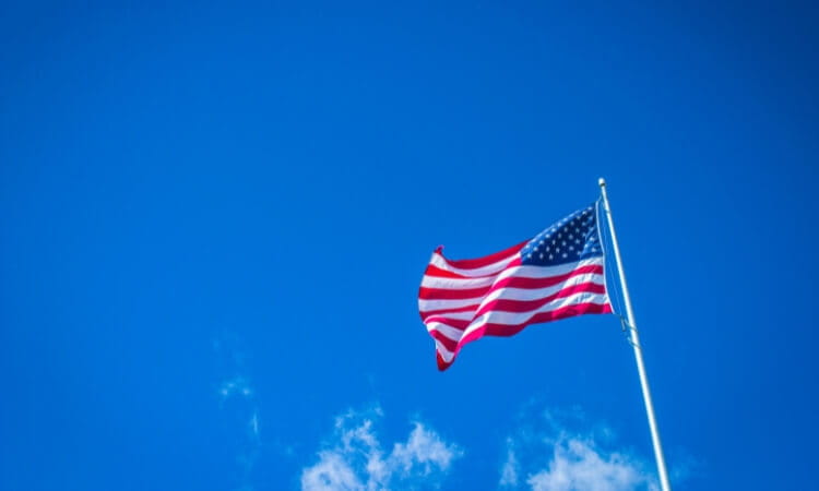 The United States flag waves on a flagpole against a clear blue sky with a few scattered clouds near the bottom.