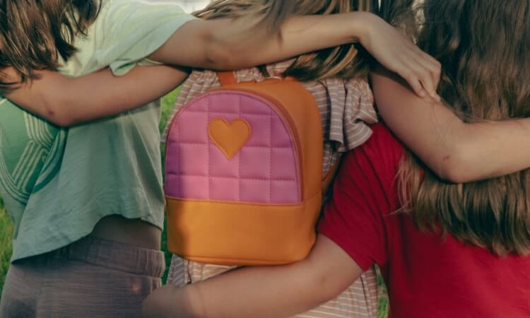 Three people standing close together with their arms around each other; one has a pink and orange backpack with a heart on it.