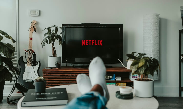 Person with feet up watching Netflix on a TV in a living room. Electric guitar and plants are in the background.