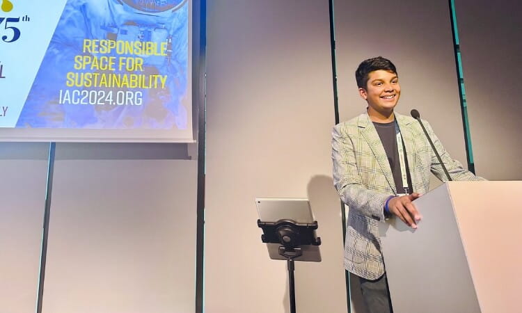 A person stands at a podium and smiles while speaking, with a screen next to them displaying "RESPONSIBLE SPACE FOR SUSTAINABILITY IAC2024.ORG.
