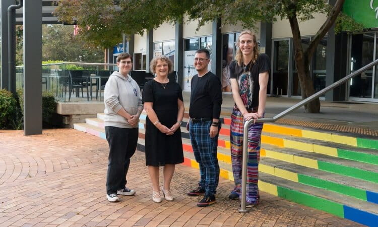Four people stand side by side outdoors on a brick path with a building and trees in the background.