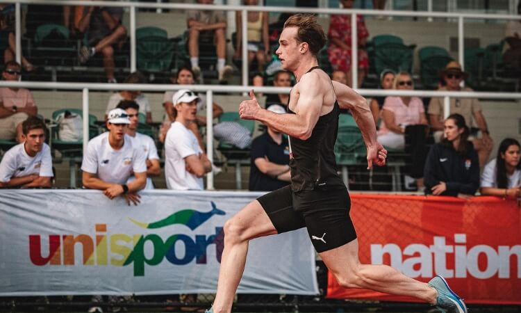 A male athlete in black athletic wear runs on a track during a race, with spectators watching from the stands in the background.