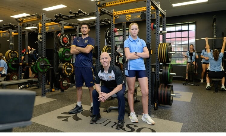Three people pose in a gym: two young athletes stand with arms crossed, while an older man kneels in front. Weightlifting equipment and other gym users are visible in the background.