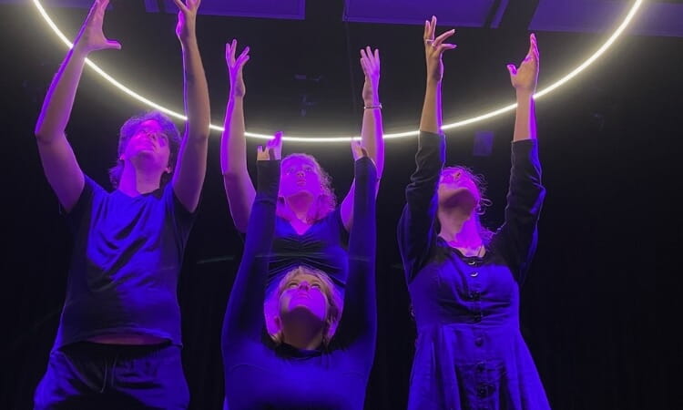 Four people in dark clothing stand under a glowing arc, reaching upward with their arms extended against a dark background, illuminated by purple lighting.