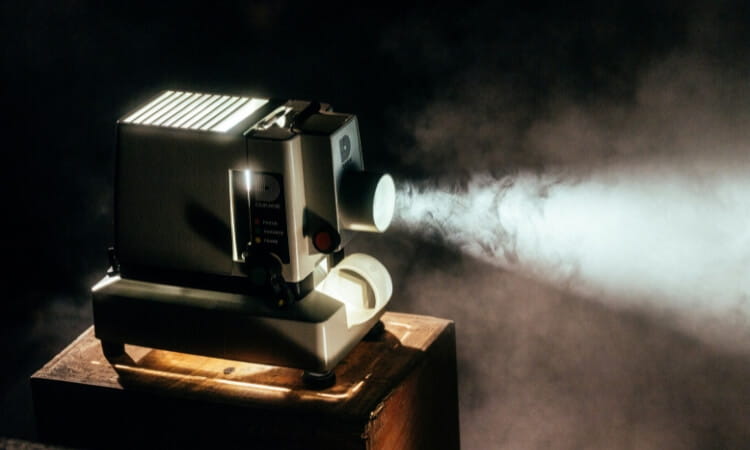 A vintage film projector sits on a wooden box, emitting a bright beam of light through a smoky, dark background.