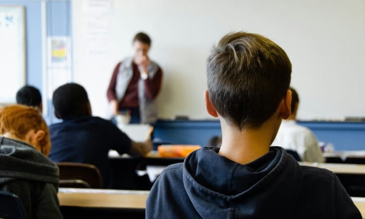 Students sit at desks, facing a teacher who stands at the front of a classroom near a whiteboard.