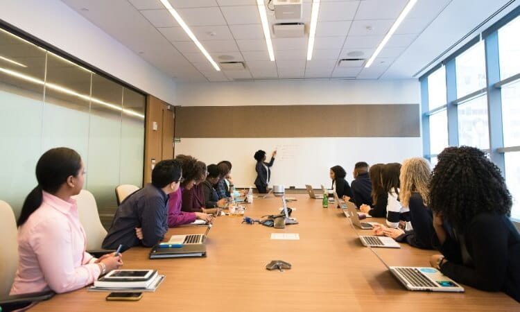 A group of people sit around a large conference table with laptops while one person stands at a whiteboard giving a presentation.