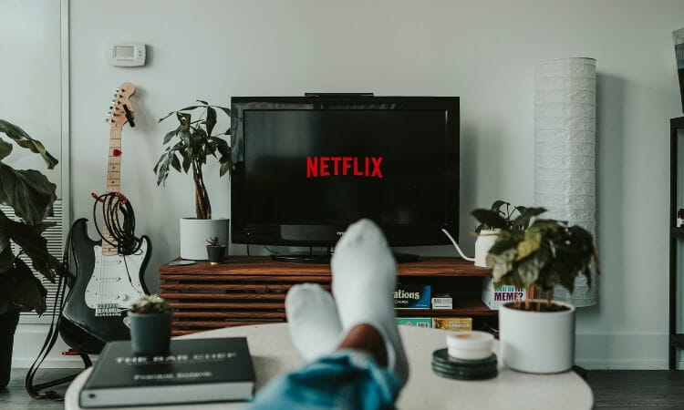 Person relaxing with feet up, watching a TV displaying the Netflix logo in a modern living room with plants and a guitar.
