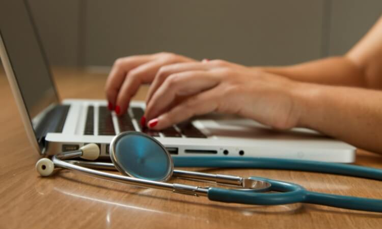 Hands typing on a laptop keyboard with a stethoscope resting on the wooden desk next to the computer.