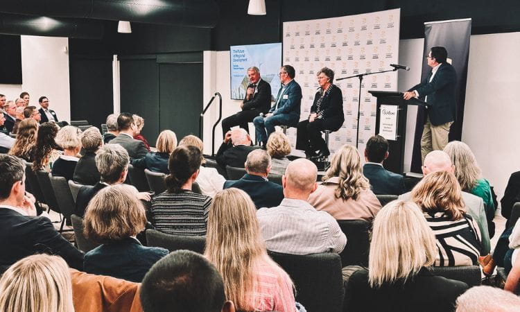 A panel of four speakers addresses a seated audience in a conference room, with one person moderating and a presentation screen visible in the background.