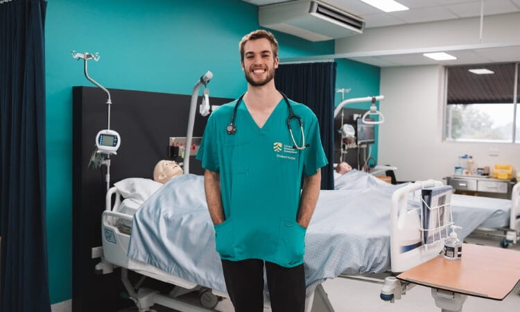 A healthcare professional in green scrubs stands smiling in a hospital room with two patient beds, one occupied by a medical mannequin.