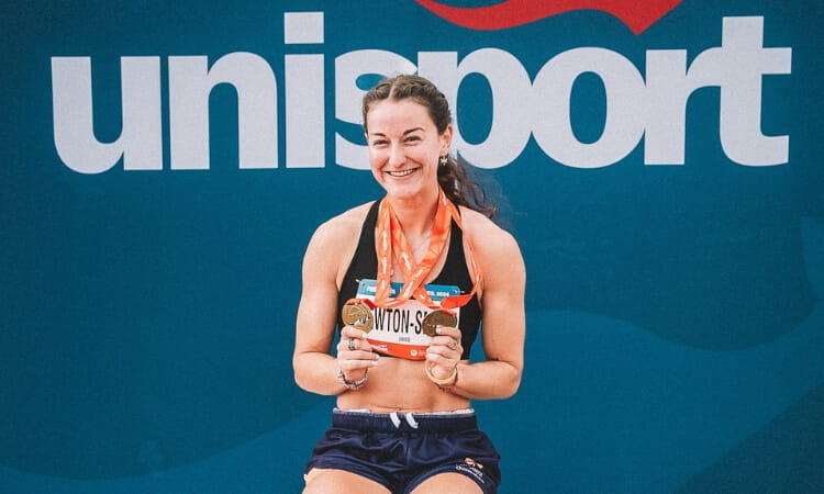 A female athlete smiles and holds up medals while sitting in front of a blue background with "UniSport" written on it.