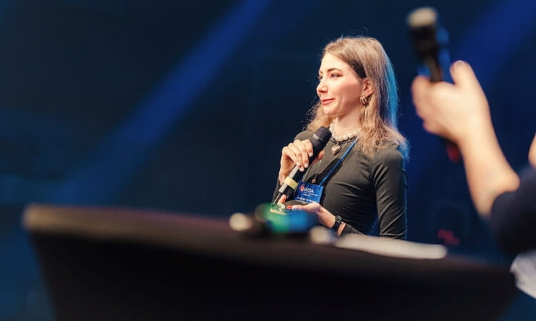 A woman holding a microphone speaks on stage at an event, with another person holding a microphone visible in the foreground.