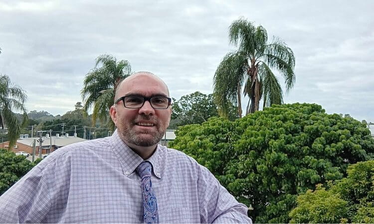 A man wearing glasses and a checkered shirt with a tie stands outdoors in front of trees and palm trees under a cloudy sky.