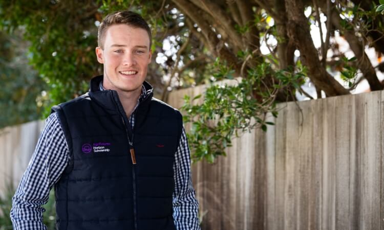 A man in a checkered shirt and dark vest stands outdoors by a wooden fence and trees, smiling at the camera.