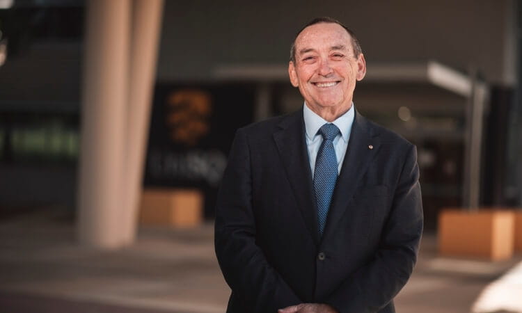A middle-aged man in a suit and tie stands smiling outdoors in front of a modern building.