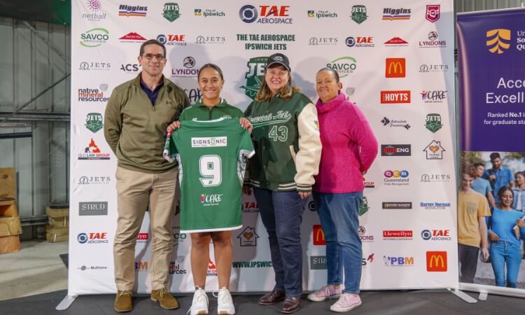 Four people stand in front of a sponsor backdrop, with one person holding a green sports jersey displaying the number 9.