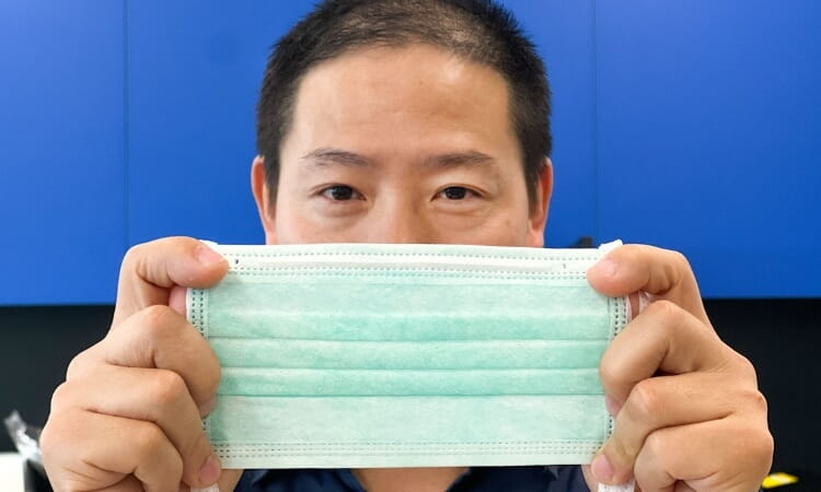 A person holds a light green disposable face mask with both hands, displaying it in front of their face against a blue and black background.