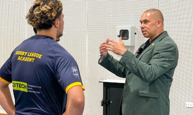 A man in a suit gestures while speaking to a person wearing a "Rugby League Academy" shirt in an indoor setting.