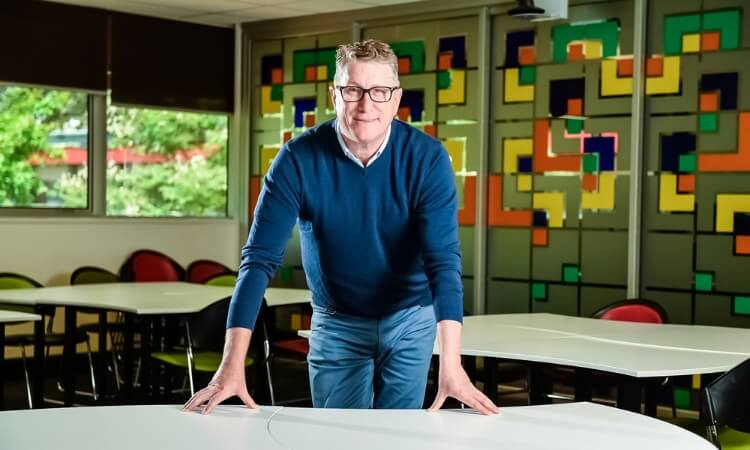 A man wearing glasses and a blue sweater stands leaning forward on a white table in a colorful classroom with geometric wall designs.