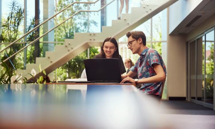 Two young adults sit at a table inside a bright building, looking at a laptop together near large windows and a staircase.