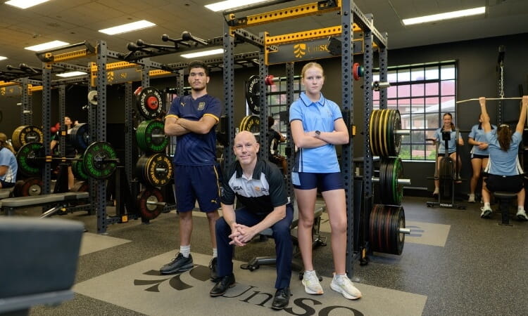 Three people pose in a gym with weightlifting equipment; two stand with arms crossed, one kneels in front. Other individuals exercise in the background.