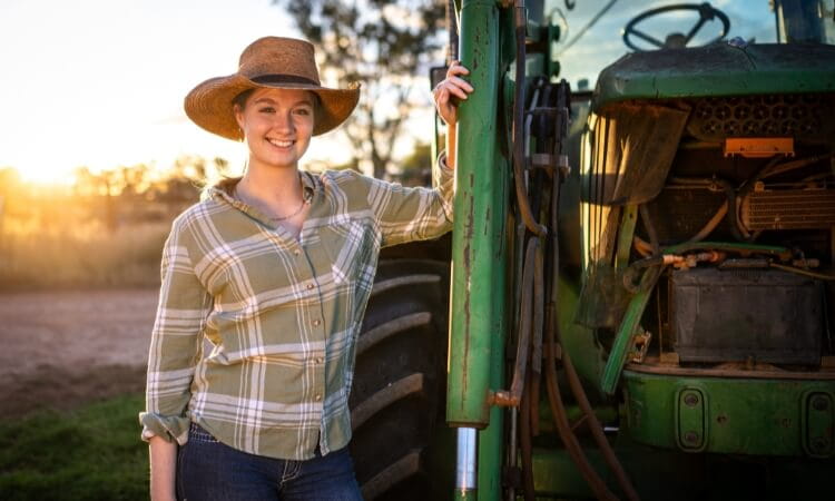 A woman wearing a plaid shirt and straw hat stands smiling beside a green tractor outdoors at sunset.