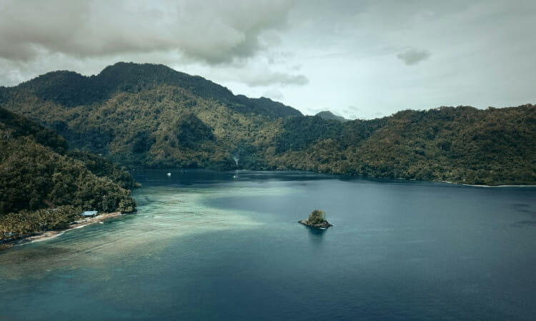 A small island sits in the middle of a calm bay surrounded by forested hills under a cloudy sky.