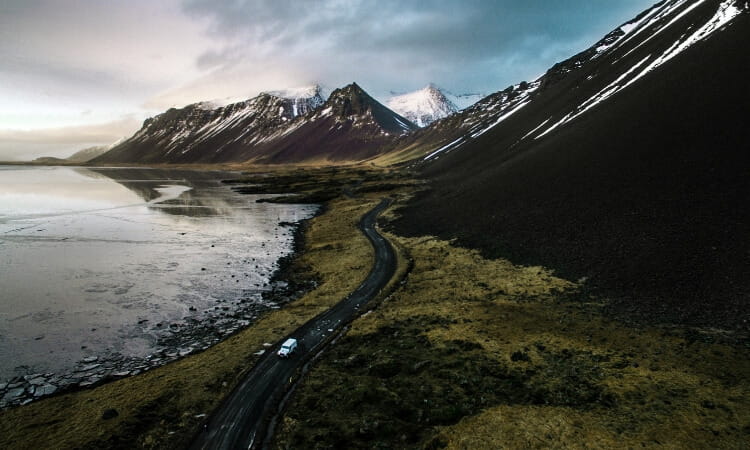 A car drives along a winding road beside a lake, with snow-capped mountains and steep slopes in the background under a cloudy sky.