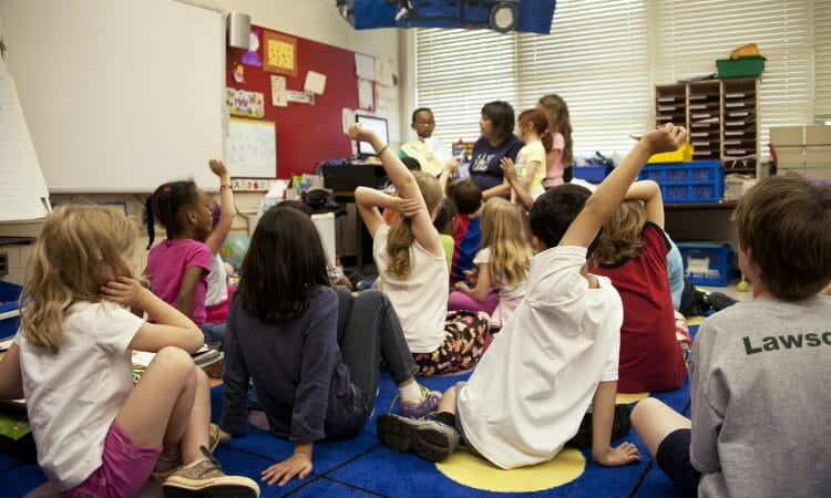 Children sit on a classroom floor, some raising their hands, while a teacher and another adult engage with them at the front near a whiteboard and red bulletin board.
