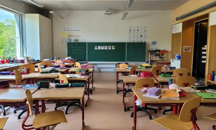A classroom with empty desks and chairs, colorful supplies on the tables, a large green chalkboard, and educational posters on the walls. Sunlight comes in through a window on the left.
