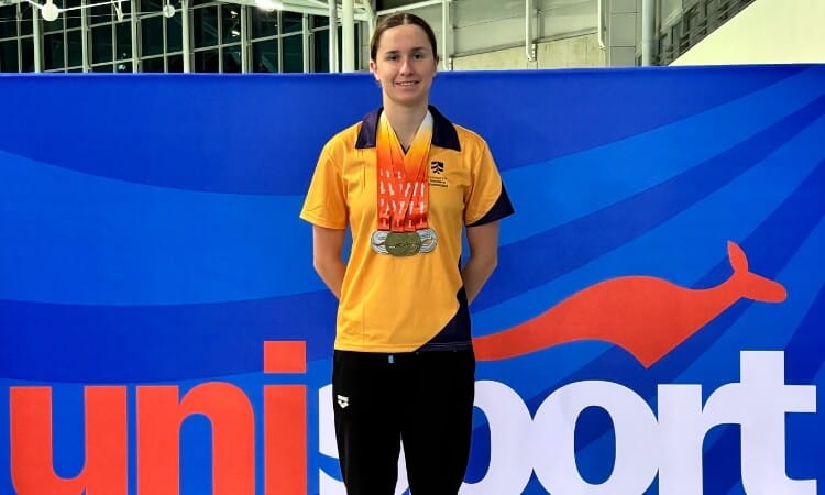 A swimmer wearing a yellow shirt and holding several medals stands in front of a blue and orange Unisport banner.