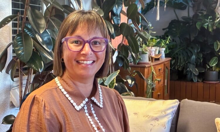 Woman with short brown hair and purple glasses sits on a sofa, smiling, with indoor plants and a wooden cabinet in the background.