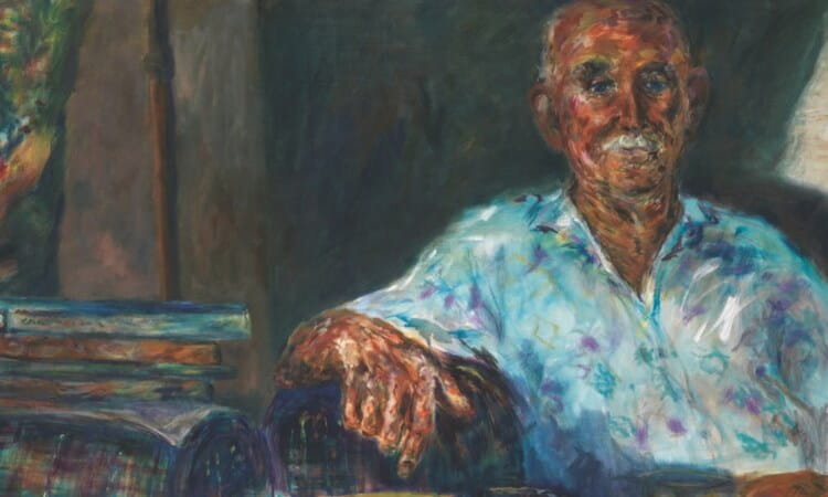 An older man with a mustache sits indoors, wearing a light-patterned shirt, with one arm resting on a stack of books.