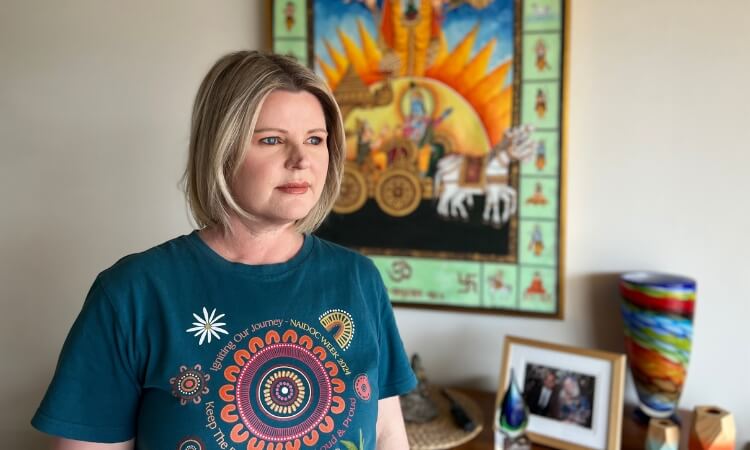 A woman with shoulder-length blonde hair stands indoors, wearing a patterned T-shirt. Behind her are a colorful painting, a framed photo, and a vibrant glass vase.