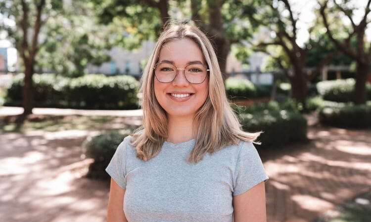 A young woman with blonde hair and glasses, wearing a grey t-shirt, stands outdoors in a park with trees and sunlight in the background.