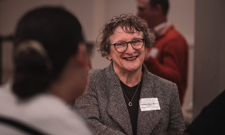 An woman smiles while talking with others at an indoor event, wearing a name tag and a grey blazer.