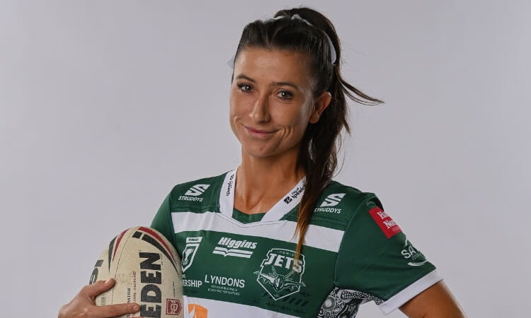 A woman in a green and white rugby jersey holds a rugby ball and stands against a plain light background.