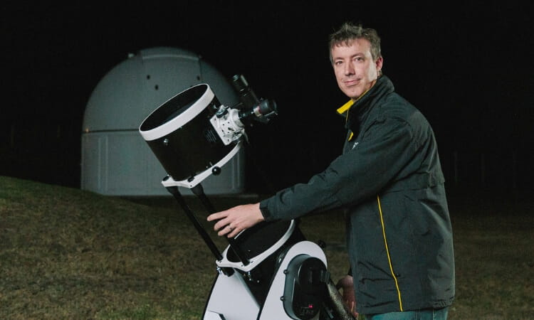 A person stands outdoors at night, adjusting a large telescope, with a white observatory dome visible in the background.
