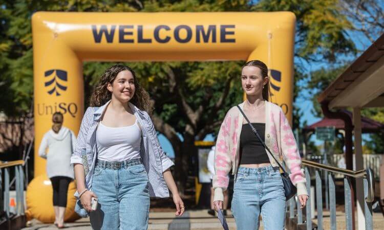 Two young women walk under a large yellow "WELCOME" arch at UniSQ, with other people in the background.