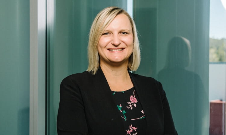A woman with blonde hair, wearing a black blazer and floral top, stands in front of a glass wall, smiling at the camera.
