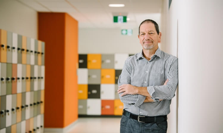 A man in a checkered shirt stands with arms crossed, leaning against a white wall in a hallway lined with colorful lockers.