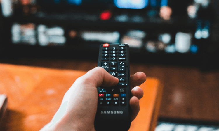 A hand holding a Samsung TV remote pointed at a television with a wooden table and shelves in the background.