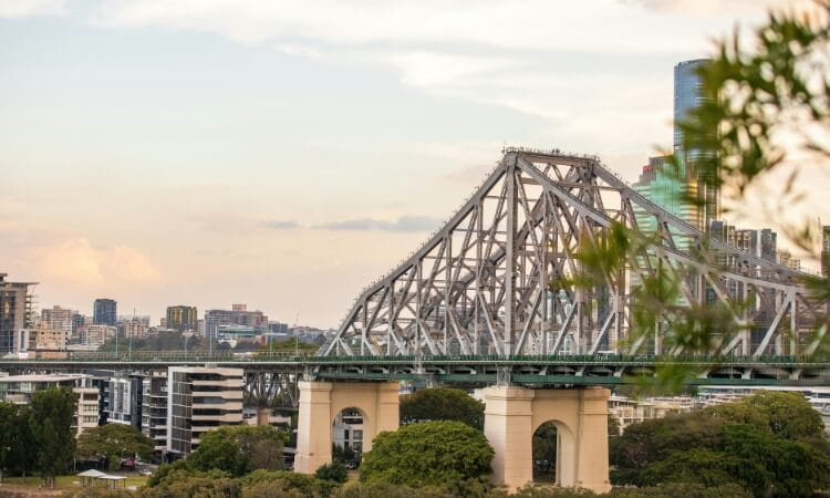 A large steel cantilever bridge spans over buildings and trees with a city skyline in the background under a partly cloudy sky.