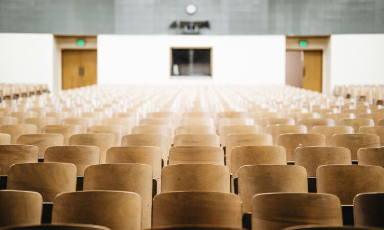 Rows of empty wooden seats in a large lecture hall facing a stage with a clock on the back wall.