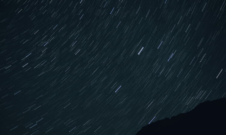 Star trails streak across the night sky above a dark, silhouetted mountain ridge, indicating a long exposure photograph capturing Earth's rotation.